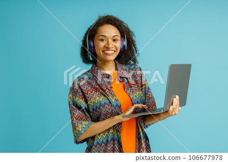 Smiling woman freelancer in headphones working on laptop while standing on blue studio background Smiling woman freelancer in headphones working on laptop while standing on blue studio background 106677978