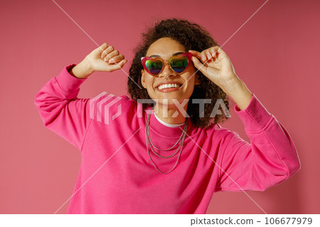 Joyful woman with curly hair is touching heart-shaped glasses on pink studio background 106677979