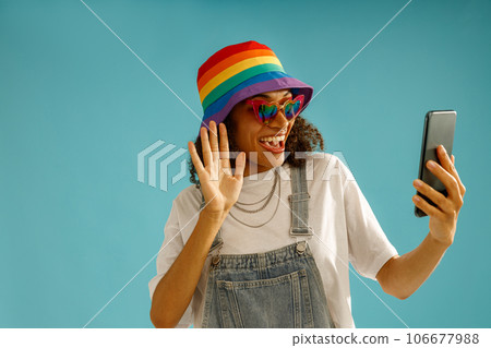 Cheerful woman in rainbow cap talking via video call and waving hi over blue studio background Cheerful woman in rainbow cap talking via video call and waving hi over blue studio background 106677988
