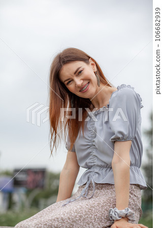 Smile of a young and beautiful girl with braces on her white teeth. A girl with long hair and a dress poses in the park. Straightening crooked teeth using a braces system. Smile of a young and beautiful girl with braces on her white teeth. A girl with long hair and a dress poses in the park. Straightening crooked teeth using a braces system. 106682939