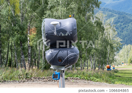Yellow snow cannon on the edge of a hill overlooking a valley in summer or warm winter, green mountain peaks. Ski slope without snow in summer or during warm winter, greenhouse effect concept. 106684865