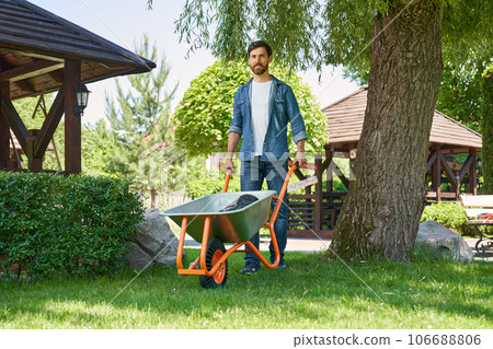 Attractive male landscaper pushing metal wheelbarrow, while working in garden. Low angle view of smiling gardener in jeans using cart for transportation, while walking on grass. Concept of gardening. 106688806