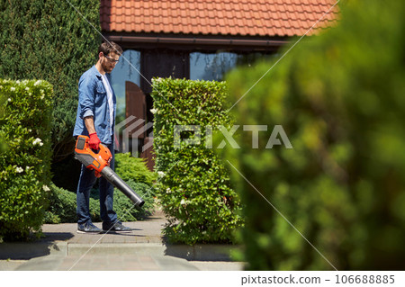 Serious mid adult man cleaning up territory with professional leaf blower in park. Side view of focused man in casual clothes holding big portable blower in early fall morning. Concept of workplace. Serious mid adult man cleaning up territory with professional leaf blower in park. Side view of focused man in casual clothes holding big portable blower in early fall morning. Concept of workplace. 106688885