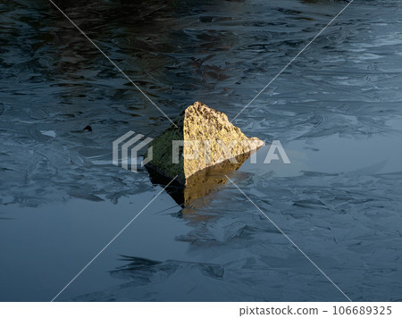 Alone rock in frozen pond. A stone that is half submerged in morning frozen water and exposed to the sunlight. 106689325