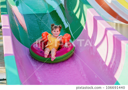 Little preschool girl on water slide in aqua park. Happy child having fun on water slides on family summer vacation in tropical resort. Amusement park with wet playground for young child and baby. 106691440