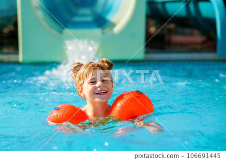 Little preschool girl with protective swimmies playing in outdoor swimming pool by sunset. Child learning to swim in outdoor pool, splashing with water, laughing and having fun. Family vacations. Little preschool girl with protective swimmies playing in outdoor swimming pool by sunset. Child learning to swim in outdoor pool, splashing with water, laughing and having fun. Family vacations. 106691445