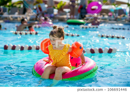 Happy little girl with inflatable toy ring float in swimming pool. Little preschool child learning to swim and dive in outdoor pool of hotel resort. Healthy sport activity and fun for children. Happy little girl with inflatable toy ring float in swimming pool. Little preschool child learning to swim and dive in outdoor pool of hotel resort. Healthy sport activity and fun for children. 106691446