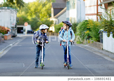 Two school kid boys in safety helmet riding with scooter in the city with backpack on sunny day. Happy children in colorful clothes biking on way to school. 106691481