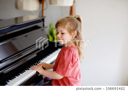Beautiful little toddler girl playing piano in living room. Cute preschool child having fun with learning to play music instrument. Early musical education for children. Beautiful little toddler girl playing piano in living room. Cute preschool child having fun with learning to play music instrument. Early musical education for children. 106691482