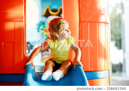 Happy blond little toddler girl having fun and sliding on indoor playground at daycare or nursery. Positive funny baby child smiling. Healthy girl climbing on slide. 106691560