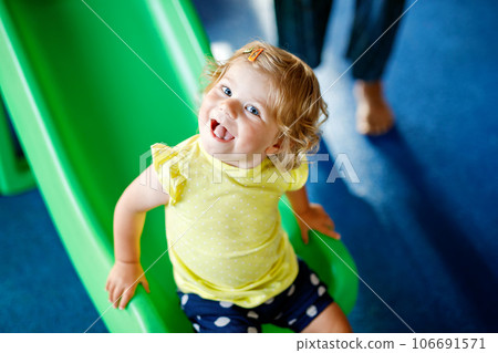 Happy blond little toddler girl having fun and sliding on indoor playground at daycare or nursery. Positive funny baby child smiling. Healthy girl climbing on slide. 106691571