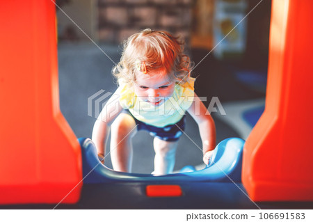 Happy blond little toddler girl having fun and sliding on indoor playground at daycare or nursery. Positive funny baby child smiling. Healthy girl climbing on slide. 106691583