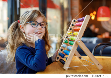 Little preschool girl playing with educational wooden rainbow toy counter abacus. Healthy happy child with glasses learning to count and colors, indoors on sunny day. Little preschool girl playing with educational wooden rainbow toy counter abacus. Healthy happy child with glasses learning to count and colors, indoors on sunny day. 106691599