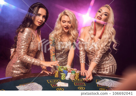 Beautiful girls with a perfect hairstyles and bright make-up are posing standing at a gambling table. Casino, poker. 106692041