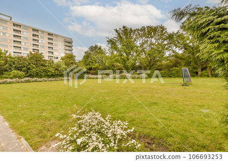 a grassy area in front of an apartment building with white flowers on the grass and trees around it, surrounded by blue sky 106693253