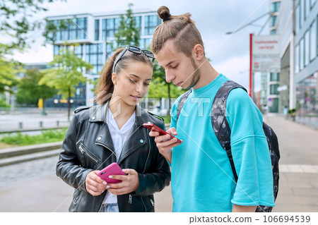 Teenage friends guy and girl standing together using smartphones Teenage friends guy and girl standing together using smartphones 106694539