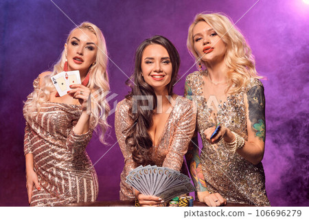 Beautiful girls with a perfect hairstyles and bright make-up are posing standing at a gambling table. Casino, poker. Beautiful girls with a perfect hairstyles and bright make-up are posing standing at a gambling table. Casino, poker. 106696279