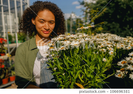 Pretty woman gardener holding flower pot while standing on greenhouse yard background 106696381