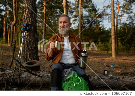 Mature man sitting drinking coffee or tea while resting in forest after hiking. 106697107