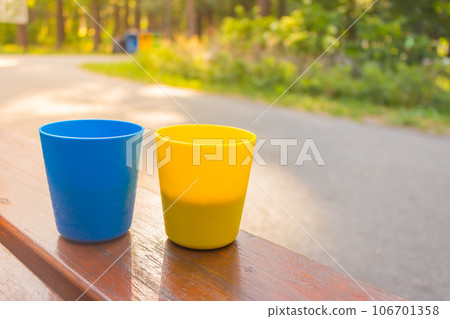 Blue and yellow cups with coffee in morning sunshine. Patriotic ukrainian colours. Two plastic mugs with cappuccino. Portable camping cups. Outdoor dating.  106701358