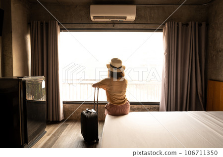 Rear view of tourist woman sitting on the bed with her luggage in hotel bedroom after check-in. Conceptual of woman lifestyle when traveling on her vacation. Rear view of tourist woman sitting on the bed with her luggage in hotel bedroom after check-in. Conceptual of woman lifestyle when traveling on her vacation. 106711350