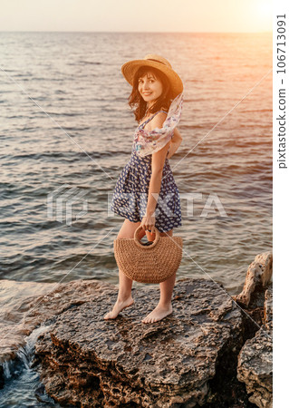 A woman in a dress, hat and with a straw bag is standing on the beach enjoying the sea. Happy summer holidays A woman in a dress, hat and with a straw bag is standing on the beach enjoying the sea. Happy summer holidays 106713091