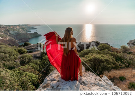 Woman sunset sea red dress, back view a happy beautiful sensual woman in a red long dress posing on a rock high above the sea on sunset. Woman sunset sea red dress, back view a happy beautiful sensual woman in a red long dress posing on a rock high above the sea on sunset. 106713092