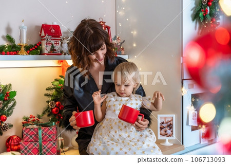 Christmas kitchen mom daughter. They are sitting on a table and drinks from a red mug in a decorated kitchen for christmas. Christmas kitchen mom daughter. They are sitting on a table and drinks from a red mug in a decorated kitchen for christmas. 106713093