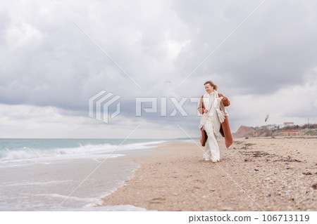 Blond woman Christmas tree sea. Christmas portrait of a happy woman walking along the beach and holding a Christmas tree on her shoulder. She is wearing a brown coat and a white suit. 106713119