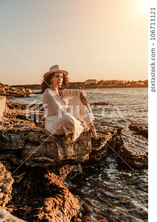 woman sea sunset. woman in a white pantsuit and hat is sitiing on the beach enjoying the sea. Happy summer holidays 106713121