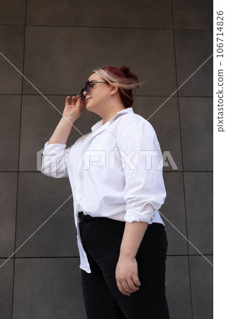 Portrait Of Fashion Plus Size Woman Peeking Over Sunglasses Outdoors. Young Stylish Female Posing Against Building Wall Background, Copy Space. Confident, Beautiful Girl. Vertical Plane. Portrait Of Fashion Plus Size Woman Peeking Over Sunglasses Outdoors. Young Stylish Female Posing Against Building Wall Background, Copy Space. Confident, Beautiful Girl. Vertical Plane. 106714826