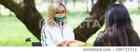 Woman with illness in wheelchair receives box with gift from her friend in park. 106715313