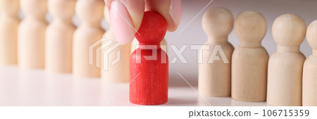 Human hand takes one red figure from group of many ordinary wooden people figures. 106715359