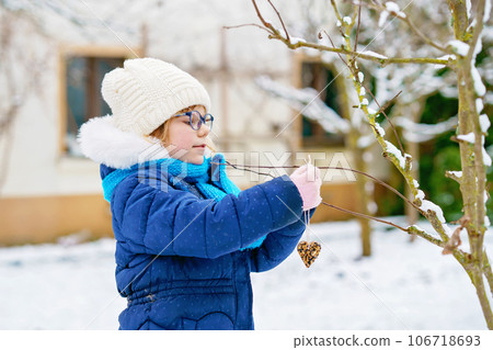 Little girl with glasses feeds birds on winter. Happy smiling preschool child hanging selfmade bird seed heart on tree. On cold snowy winter day. 106718693