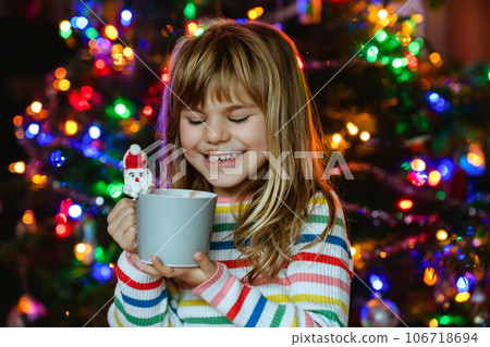 Little child girl holding cup with hot chocolate with marshmallows as Santa Claus. Kid sitting near Christmas tree decorated with lights. Little child girl holding cup with hot chocolate with marshmallows as Santa Claus. Kid sitting near Christmas tree decorated with lights. 106718694