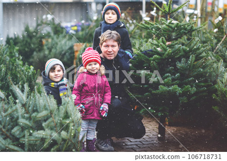 Little toddler girl, two kids boys and father holding Christmas tree on market. Happy family, cute children and middle aged man in winter fashion clothes choosing and buying Xmas tree in outdoor shop. 106718731