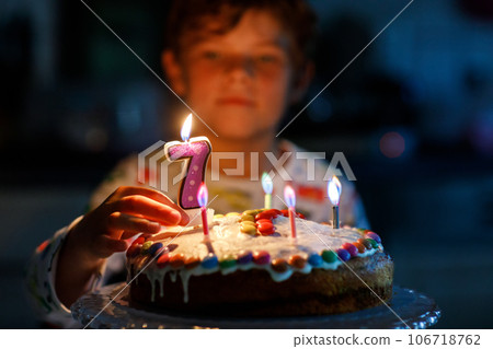 Adorable happy blond little kid boy celebrating his birthday. Child blowing seven candles on homemade baked cake, indoor. Birthday party for school children, family celebration of 7 years Adorable happy blond little kid boy celebrating his birthday. Child blowing seven candles on homemade baked cake, indoor. Birthday party for school children, family celebration of 7 years 106718762