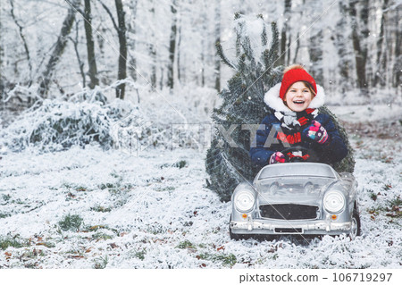 Happy little smiling girl driving toy car with Christmas tree. Funny preschool child in winter clothes bringing hewed xmas tree from snowy forest. Family, tradition, holiday. 106719297