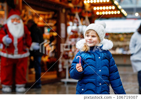 Little cute preschool girl with candy cane from a sweets stand on Christmas market. Happy child on traditional family market in Germany. Preschooler in colorful winter clothes during snowfall Little cute preschool girl with candy cane from a sweets stand on Christmas market. Happy child on traditional family market in Germany. Preschooler in colorful winter clothes during snowfall 106719307