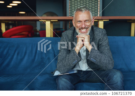 Portrait of smiling senior businessman sitting on sofa in office and looking at camera Portrait of smiling senior businessman sitting on sofa in office and looking at camera 106721370