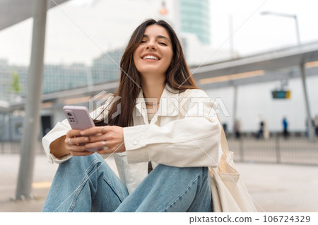 Waiting for the bus and for love. Bright cheerful young woman of Turkish mixed race in white casual clothes with a charming smile holding a phone in the street at public transportation stop Waiting for the bus and for love. Bright cheerful young woman of Turkish mixed race in white casual clothes with a charming smile holding a phone in the street at public transportation stop 106724329