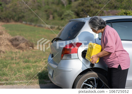 The car ran out of gas and stalled beside the road in suburbs and an elderly Asian woman used a gallon of spare gas to fuel the car. A woman prepares a gallon of spare gas to fuel before traveling. The car ran out of gas and stalled beside the road in suburbs and an elderly Asian woman used a gallon of spare gas to fuel the car. A woman prepares a gallon of spare gas to fuel before traveling. 106725052