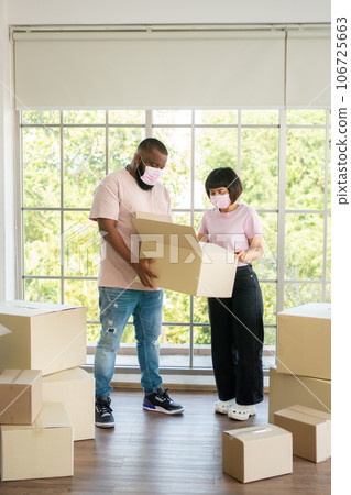 Mixed Race couple American African and Asian a surgical mask is carrying cardboard boxes in a new house on moving day. Concept of relocation, rental, and homeowner moving at home. Mixed Race couple American African and Asian a surgical mask is carrying cardboard boxes in a new house on moving day. Concept of relocation, rental, and homeowner moving at home. 106725663