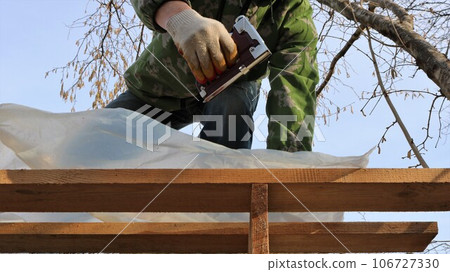 A worker in gloves holds a construction stapler and attaches plastic thick film to a wooden deck on the roof of an outbuilding to protect against moisture, attaching plastic protective material A worker in gloves holds a construction stapler and attaches plastic thick film to a wooden deck on the roof of an outbuilding to protect against moisture, attaching plastic protective material 106727330