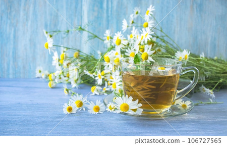 Tea from fresh chamomile flowers, mint, in a cup, on a wooden table, against the background of bouquets of fresh chamomile flowers Tea from fresh chamomile flowers, mint, in a cup, on a wooden table, against the background of bouquets of fresh chamomile flowers 106727565