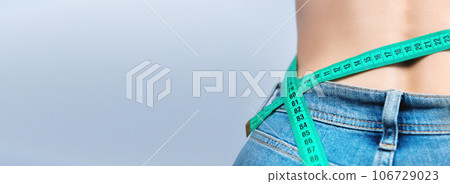 slender woman measures her waist with a tape measure. Healthy body weight loss concept. Slim waist in denim trousers, on a blue background. Long web banner slender woman measures her waist with a tape measure. Healthy body weight loss concept. Slim waist in denim trousers, on a blue background. Long web banner 106729023