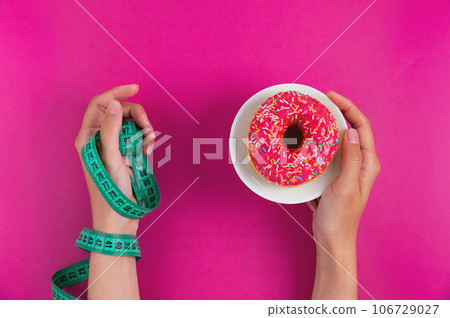 woman's hand holds a sweet donut and a measuring tape, on a pink background. Top view, flat lay. Weight loss and choice 106729027
