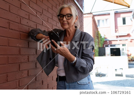 well-groomed senior woman with gray hair and glasses is dressed in a gray jacket and jeans chatting well-groomed senior woman with gray hair and glasses is dressed in a gray jacket and jeans chatting 106729161