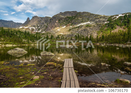 Wooden pier going into the water. Lake Radughnoe in Ergaki on an summer among the taiga rocks with blue dramatic sky and cedar trees Mountain landscape. Ergaki Nature Park in Siberia. 106730170