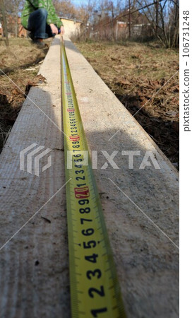 vertical shot with an example of using a tape measure in centimeters when measuring the length of a long wooden board outdoors with the beginning of a line with numbers in the foreground 106731248